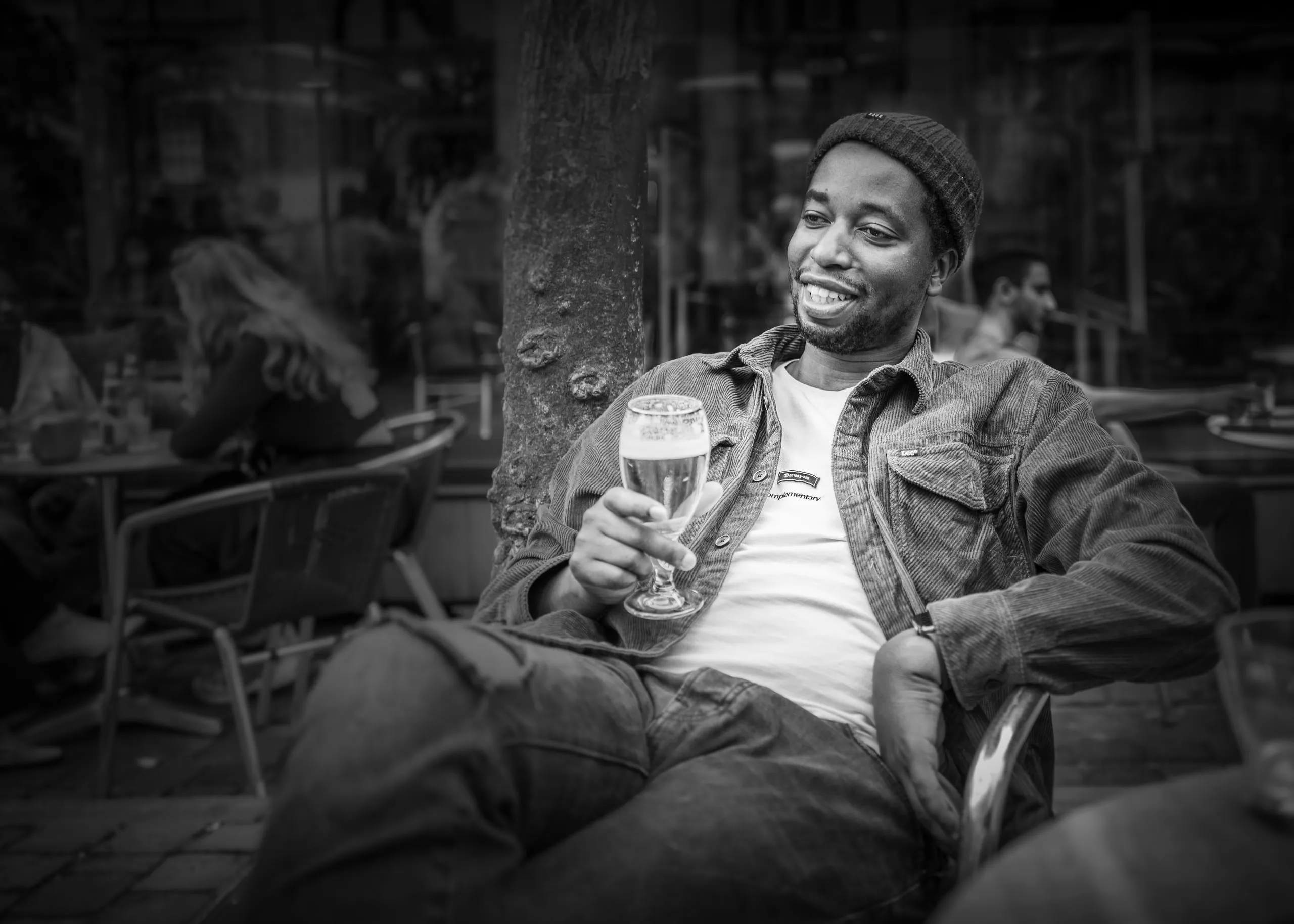 Man in a beanie laughing on a café terrace, holding a glass of beer, in black and white