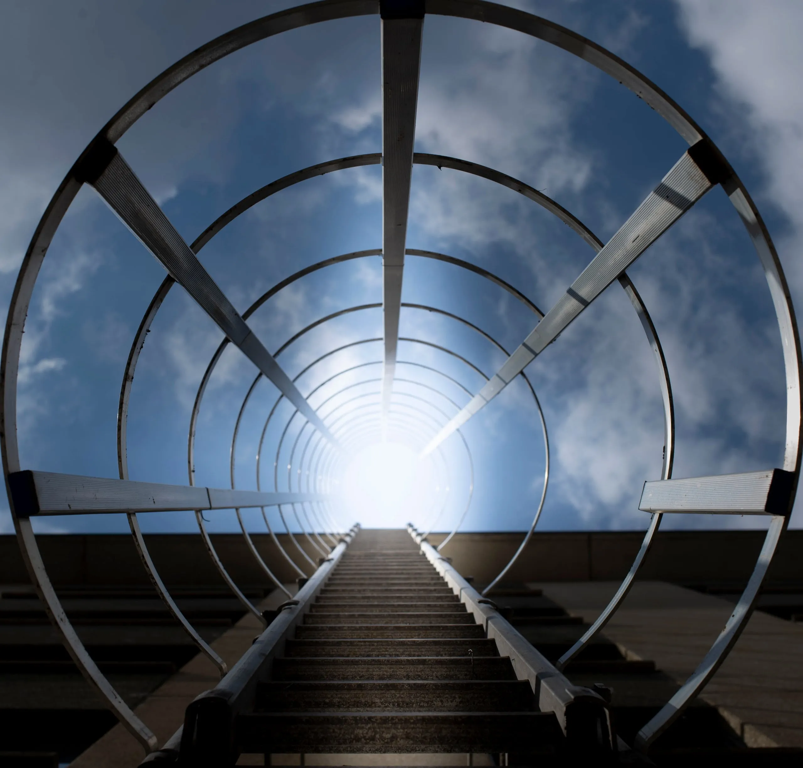 Looking up through a round metal stair cage to a blue sky with clouds
