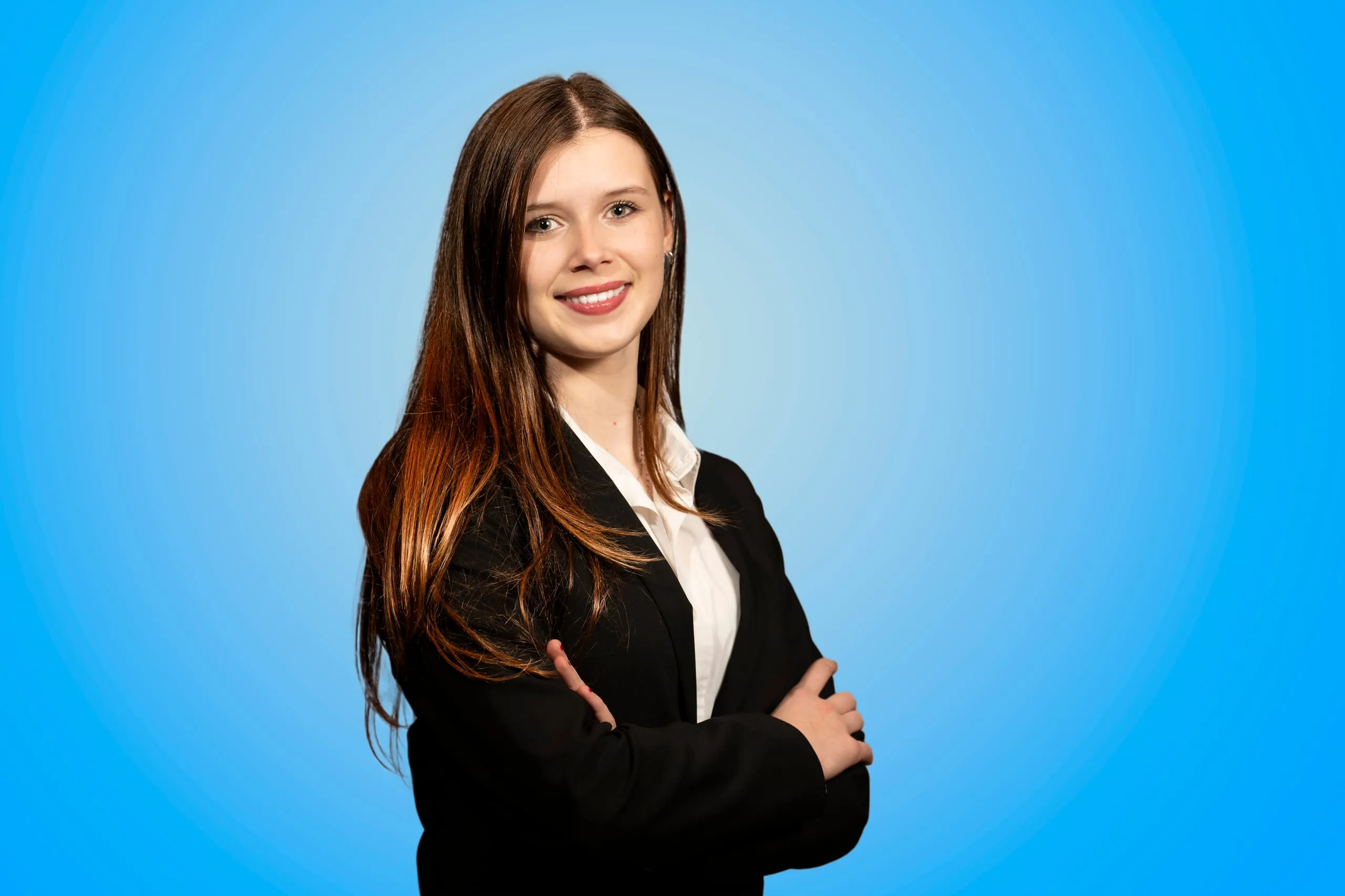 Corporate portrait of a young woman in a black blazer with her arms crossed against a blue background
