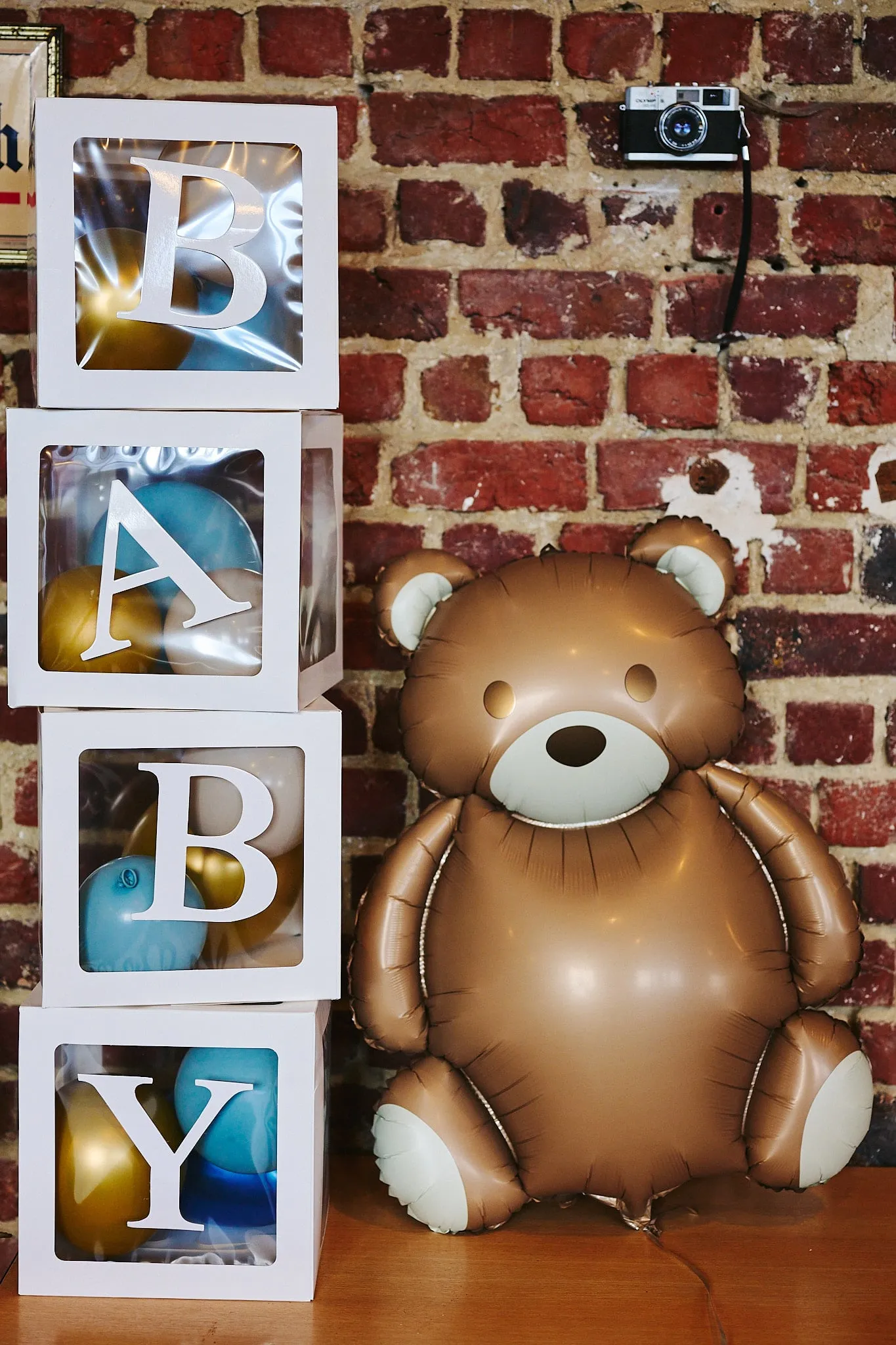 White letter blocks spelling BABY next to a large teddy bear balloon against a brick wall
