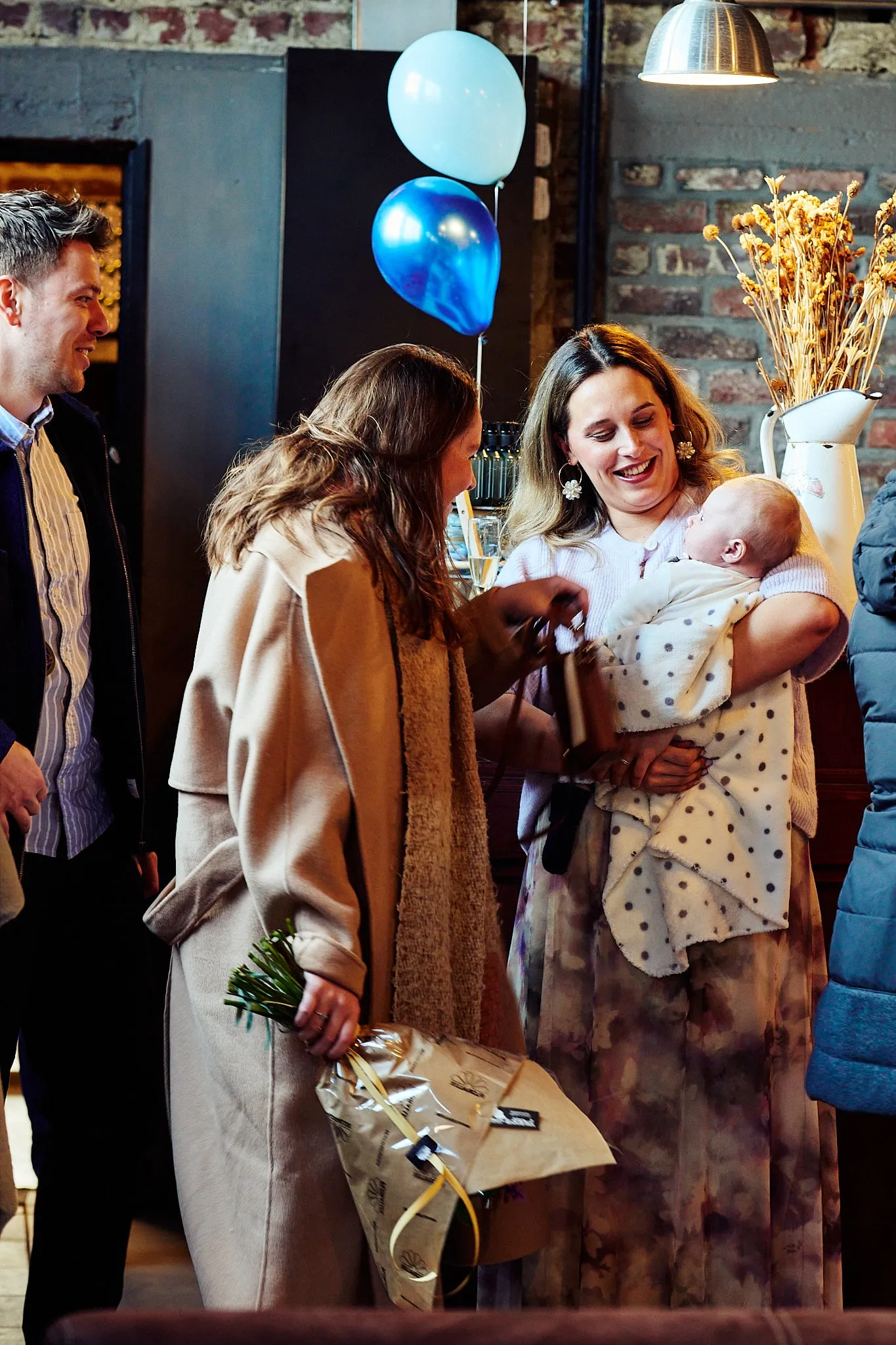 Mother with a baby in her arms greets guests, with balloons in the background