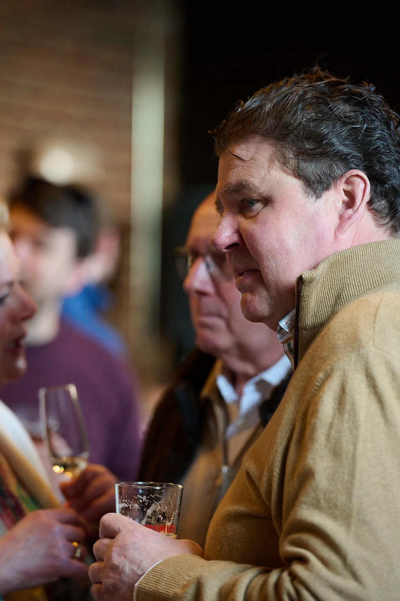 Man in a beige sweater with a glass in his hand among other guests
