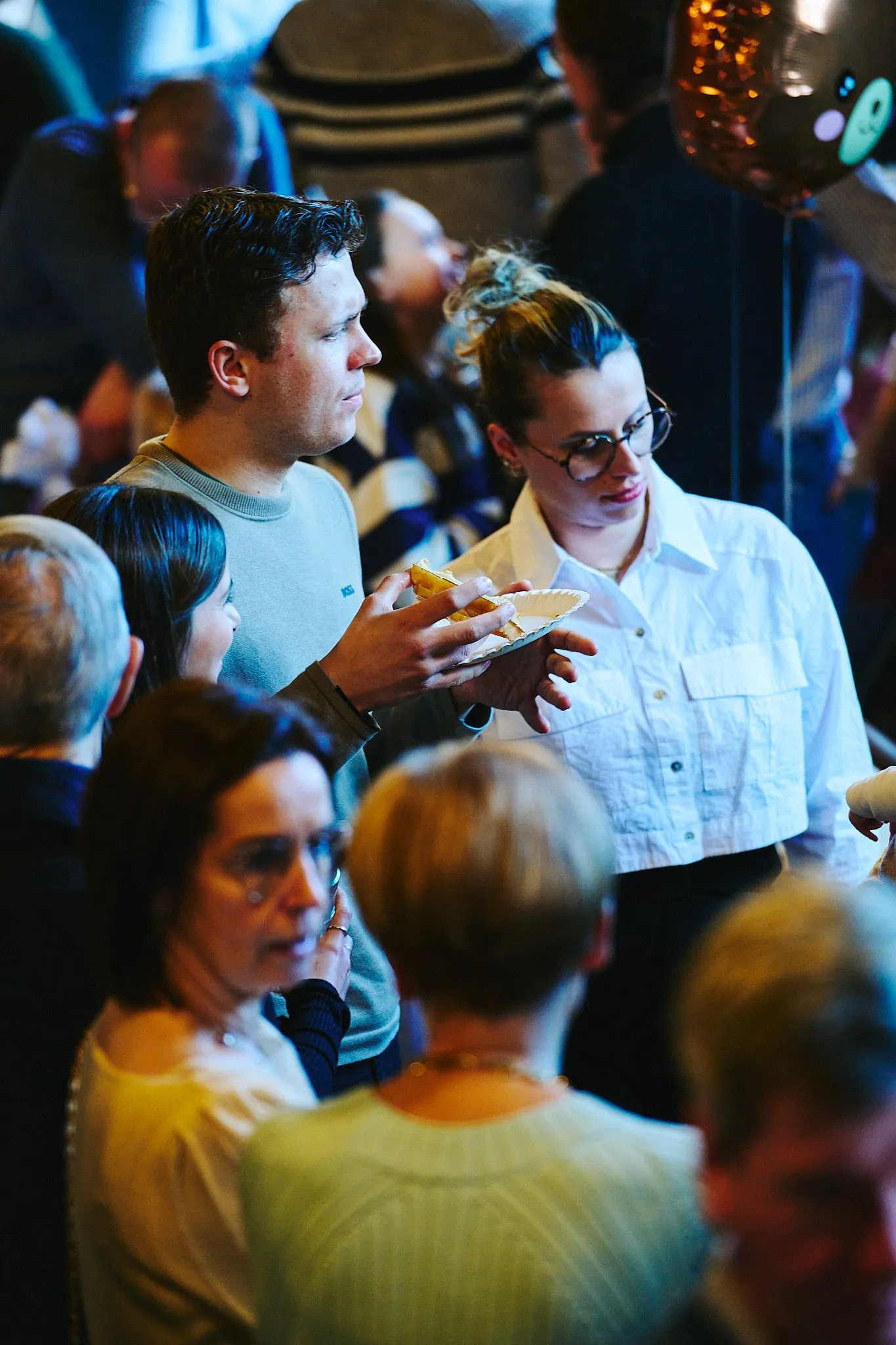 Guests in a crowded room, a woman in a white blouse holding a small plate
