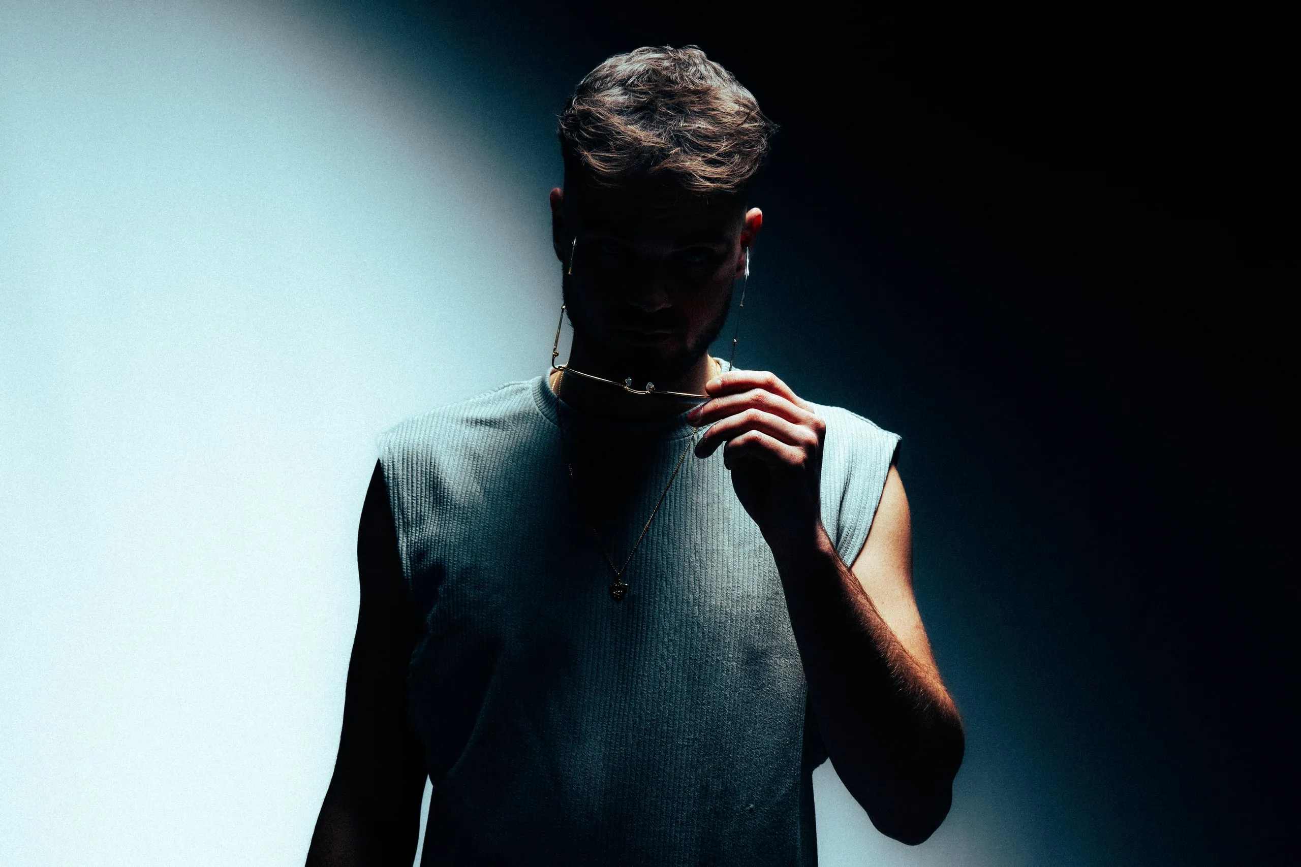 Man in a sleeveless shirt holding a glasses chain, his face in shadow against a light and dark background