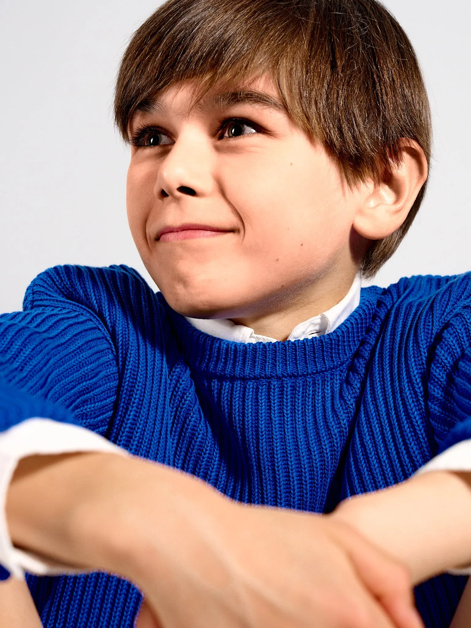Boy in a blue sweater smiling sideways at the camera, with his arms on his knees
