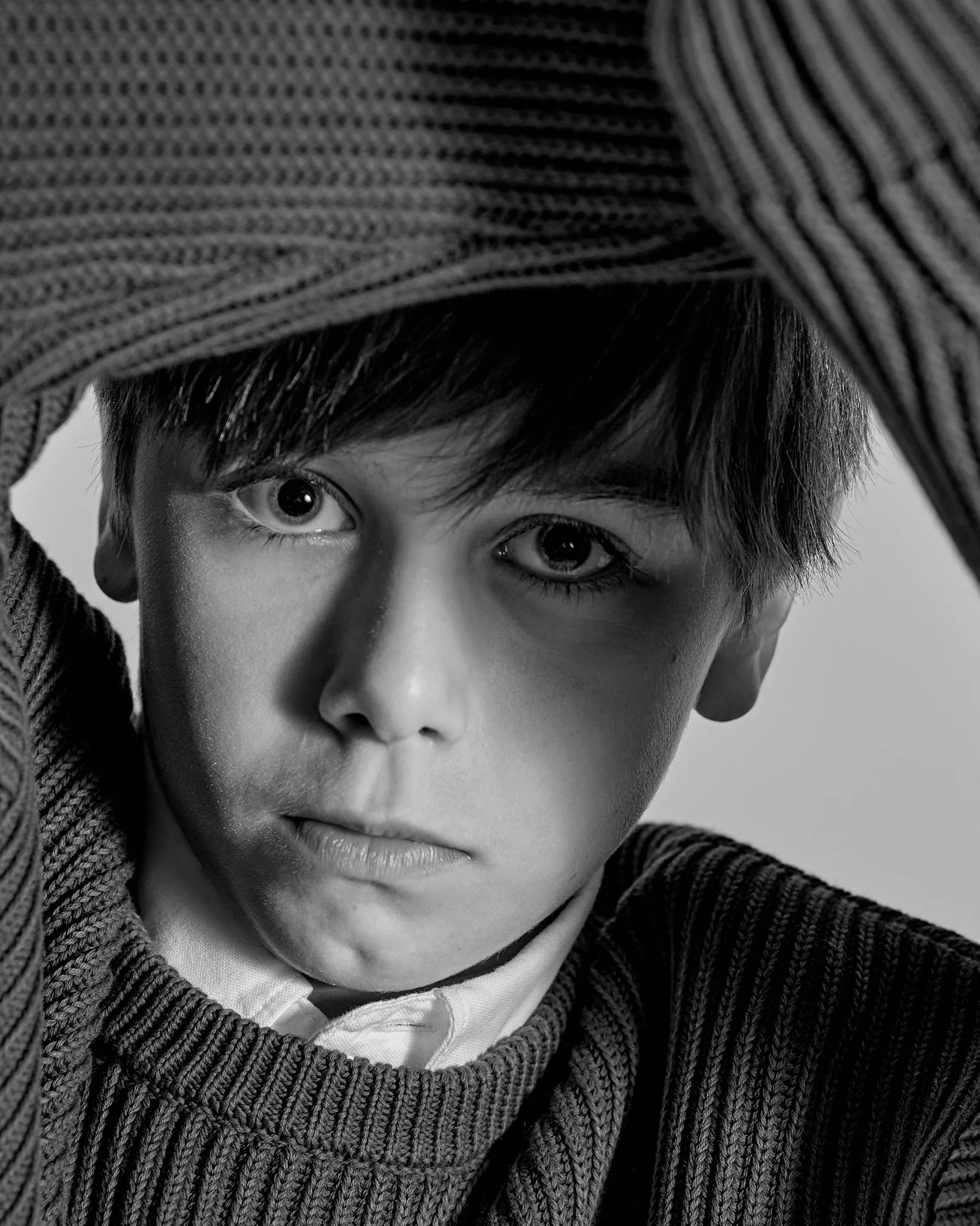 Black-and-white portrait of a boy pulling a knitted sweater over his head