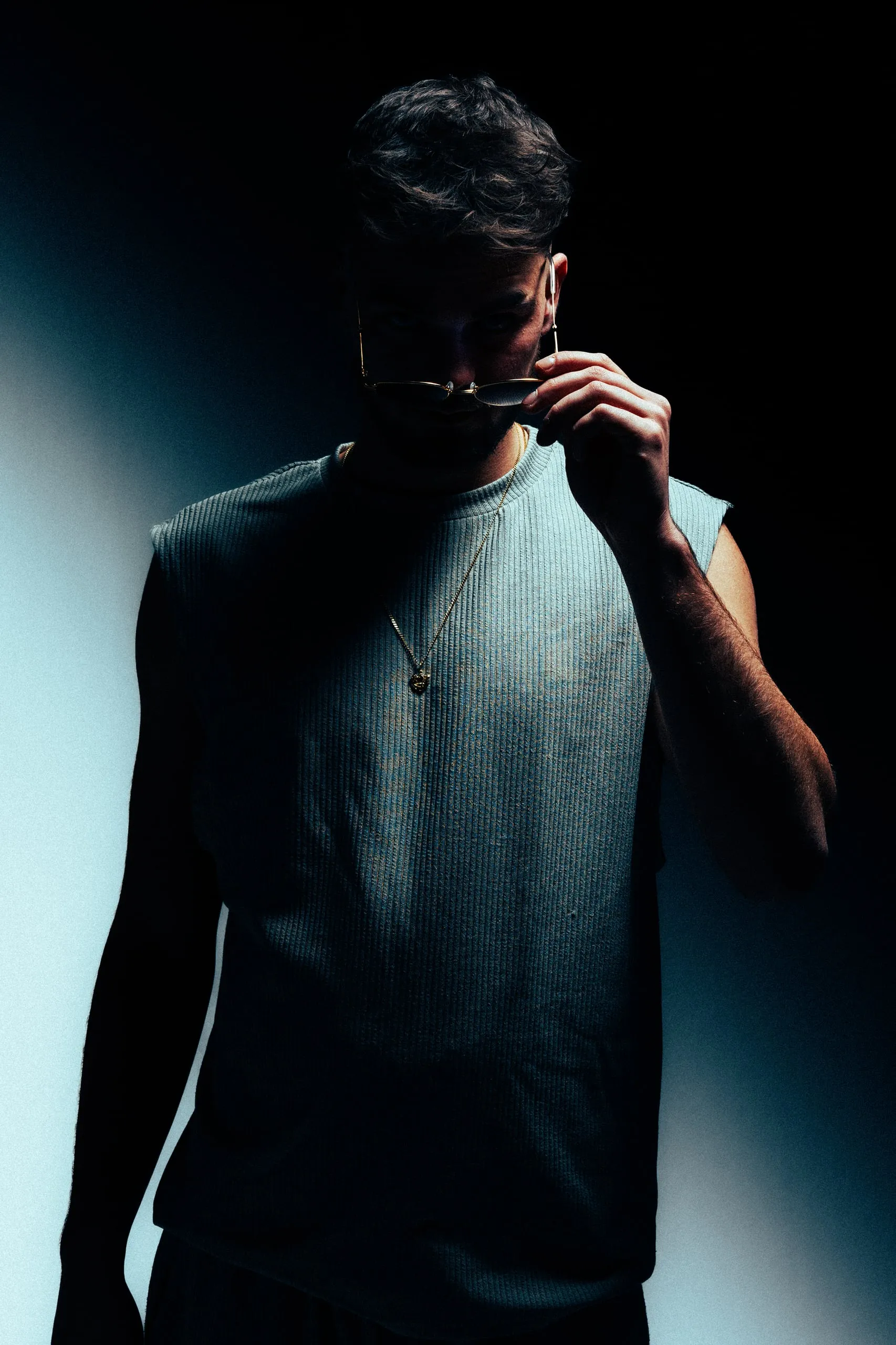 Man in a sleeveless shirt holds glasses to his lips, with sharp light and deep shadows