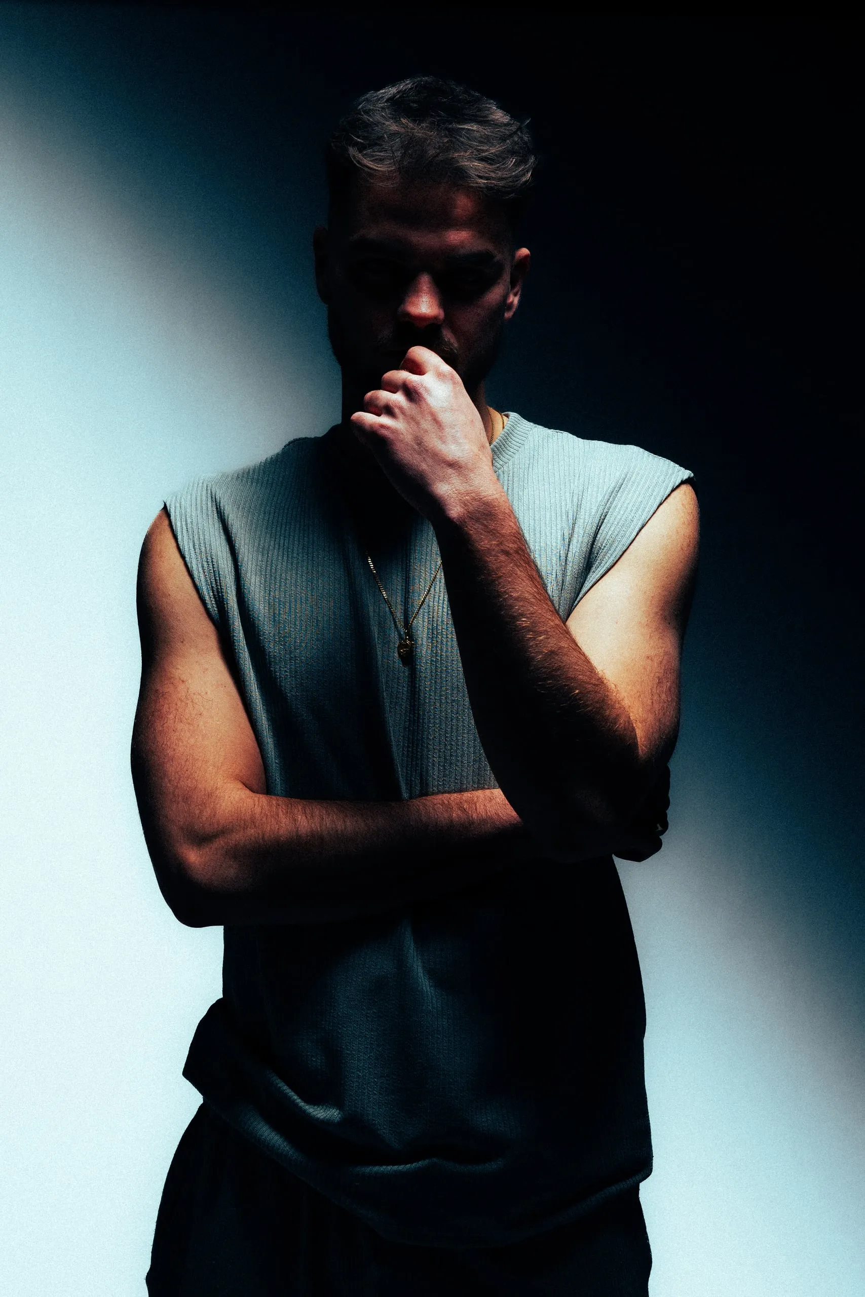 Man in a sleeveless shirt with crossed arms and a hand on his chin, his face in shadow